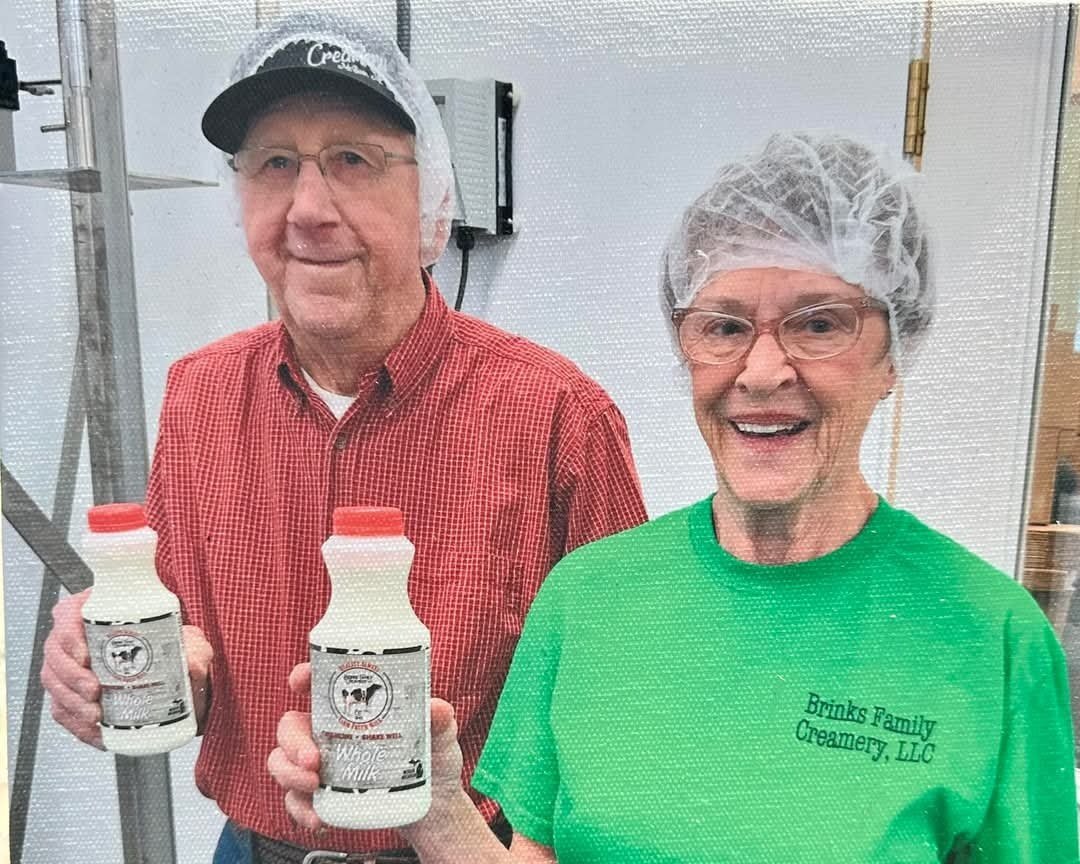 Elderly couple holding bottles of whole milk in a creamery setting, wearing hairnets and branded apparel from Brinks Family Creamery, LLC. The image showcases their commitment to dairy production and local farming.